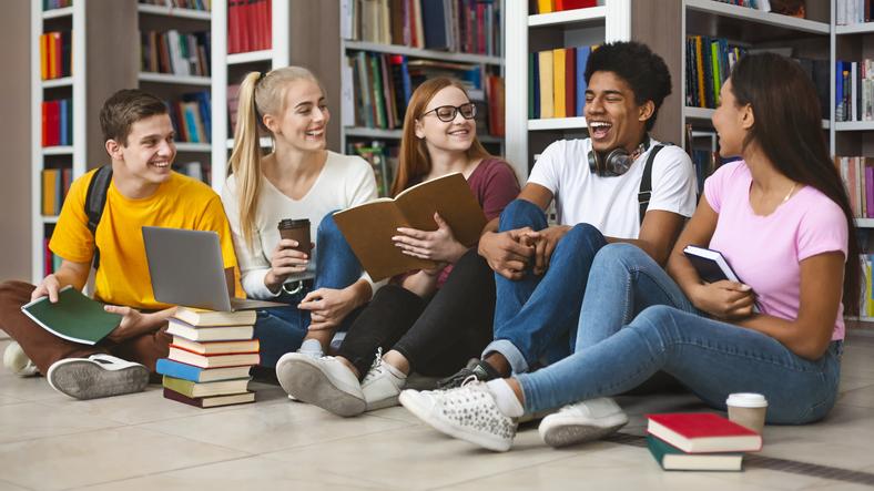 A group of students laugh together while sitting on the floor of a library