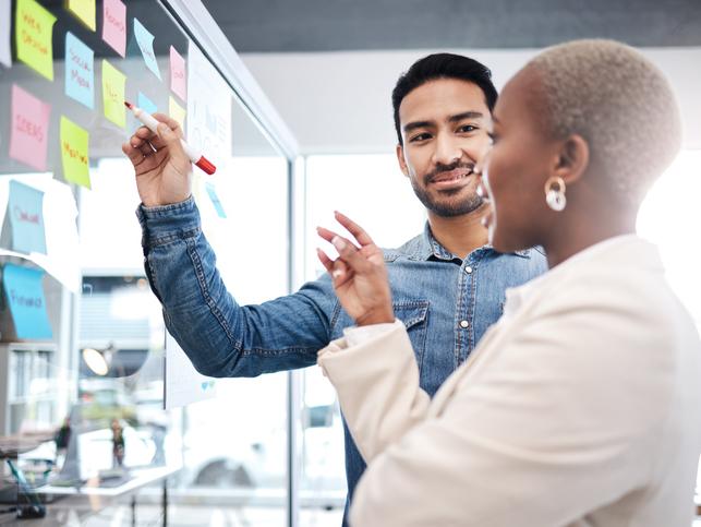 Two people gesture in front of a board covered in post-its