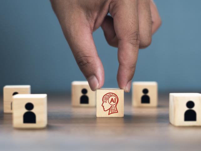 A hand reaches for a wooden block displaying "AI"