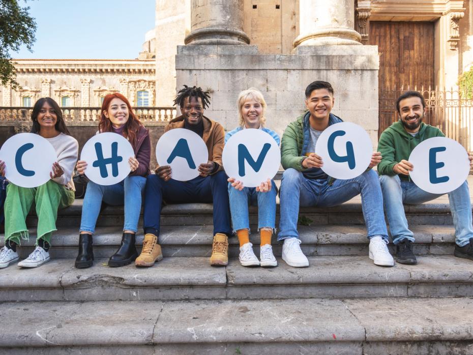 Students sitting on the steps of a campus building each holding a paper with a letter of the word "change" on it