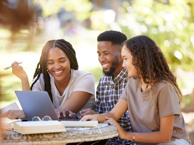 A group of students sit happily around a laptop in the sunshine