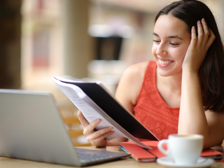 A young woman referring to her notes while stufying at home on her laptop