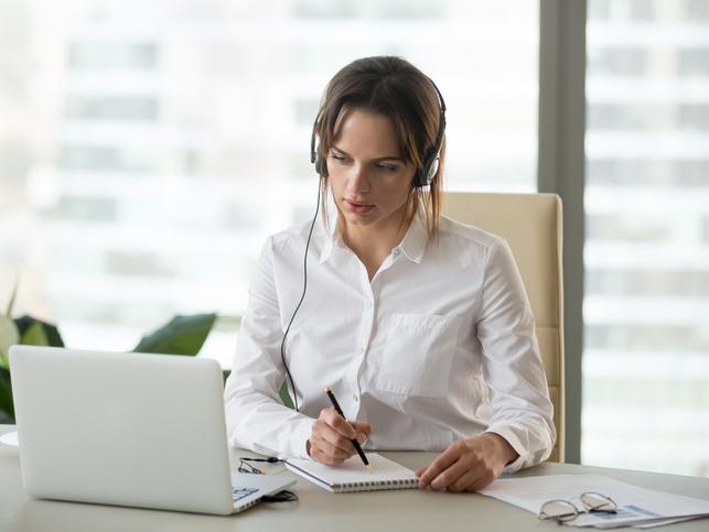 A woman watches a laptop while wearing headphones and making notes