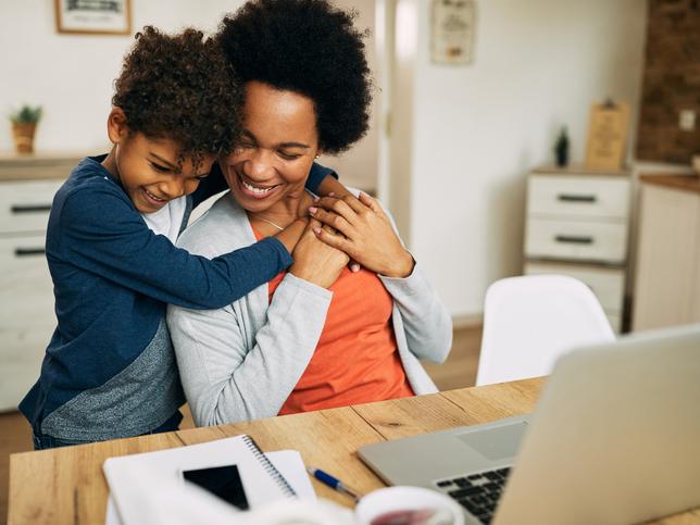 A son hugs his mother as she works at a laptop