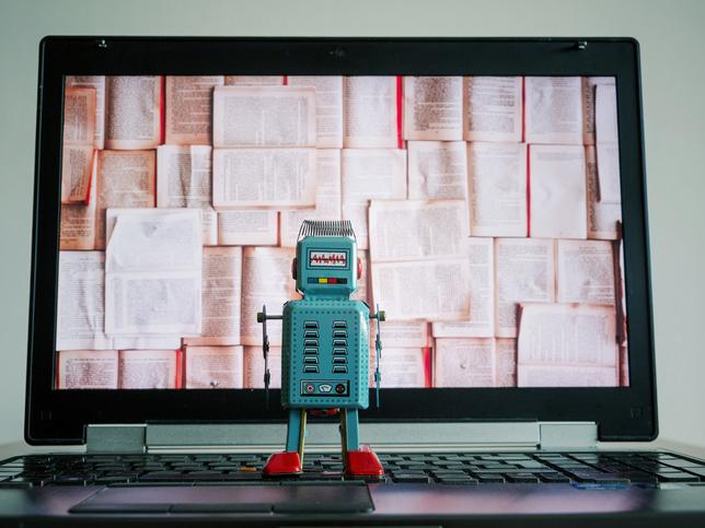 A toy robot observes a laptop screen, filled with books