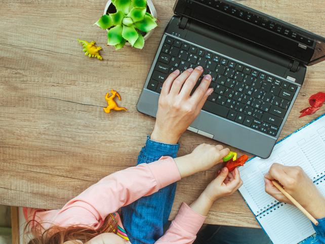 A adult works at a laptop, while a small child plays with dinosaur toys on the keyboard