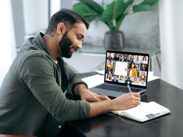 A man takes notes as he watches an online lecture on his laptop