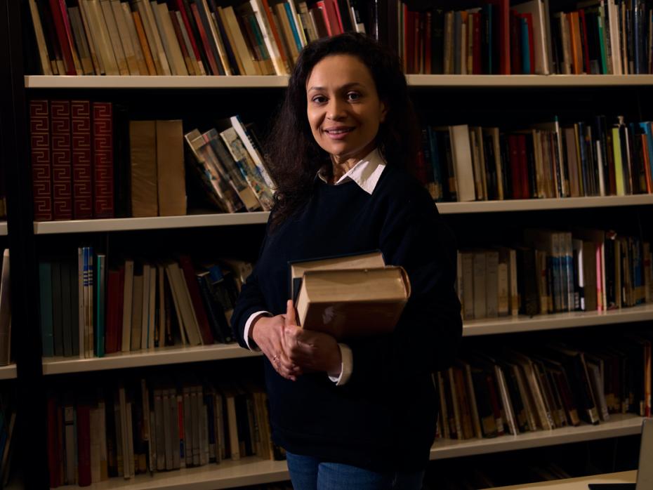 An academic holding a pile of books in front of a library bookshelf