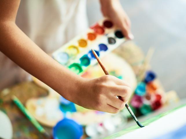 A close-up of a hand painting on a canvas, a paint palette in the background