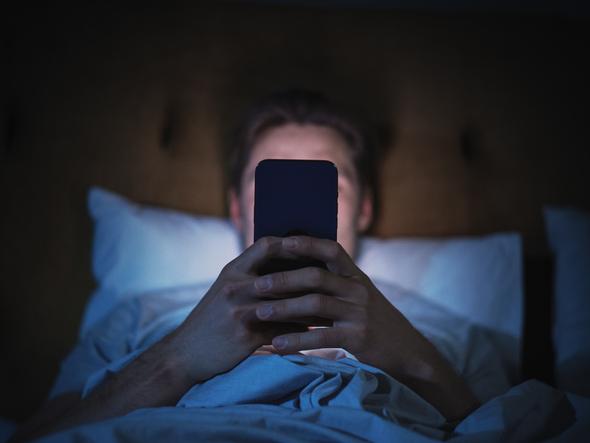 A man sits in darkness, illuminated by a smartphone that blocks his face from view