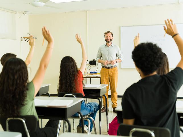A teacher smiles as students raise their hands en masse