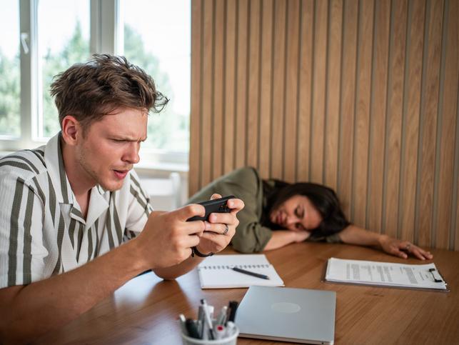 A young academic plays a game on his phone while his co-worker naps on the desk