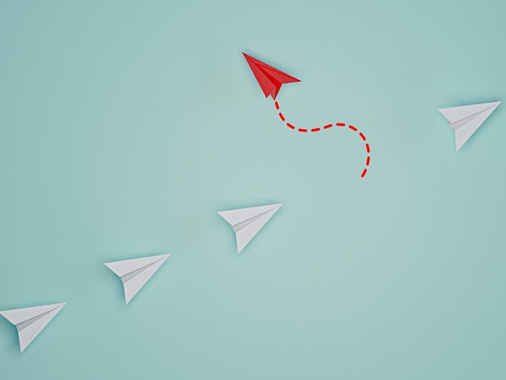 red paper airplane leaving line of white paper airplanes