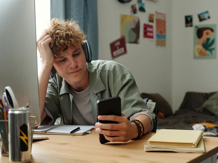 Young male student looking at phone while putting off study