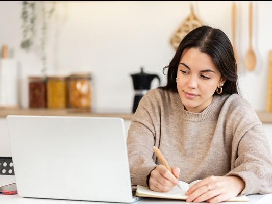 A student taking notes on a notepad while looking at her laptop