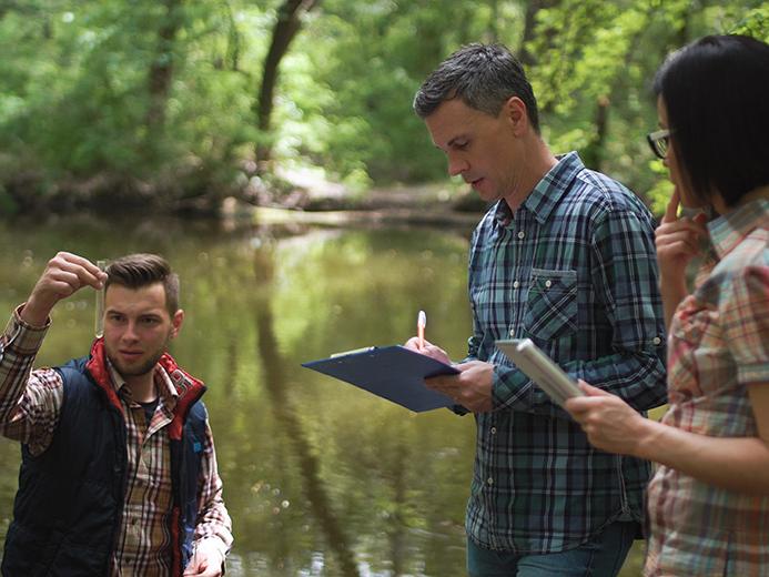 Three researchers in casual clothing at a lake take water sample