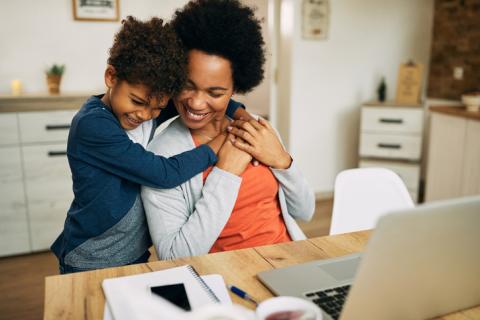 A son hugs his mother as she works at a laptop