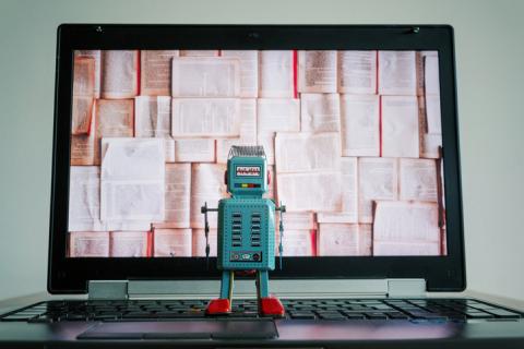 A toy robot observes a laptop screen, filled with books