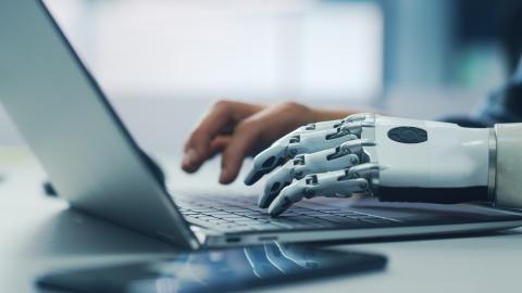 A human and robot hand typing on a laptop