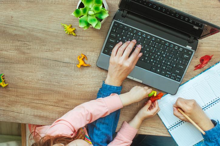 A adult works at a laptop, while a small child plays with dinosaur toys on the keyboard