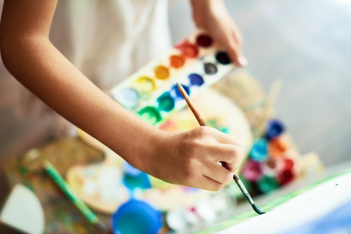 A close-up of a hand painting on a canvas, a paint palette in the background