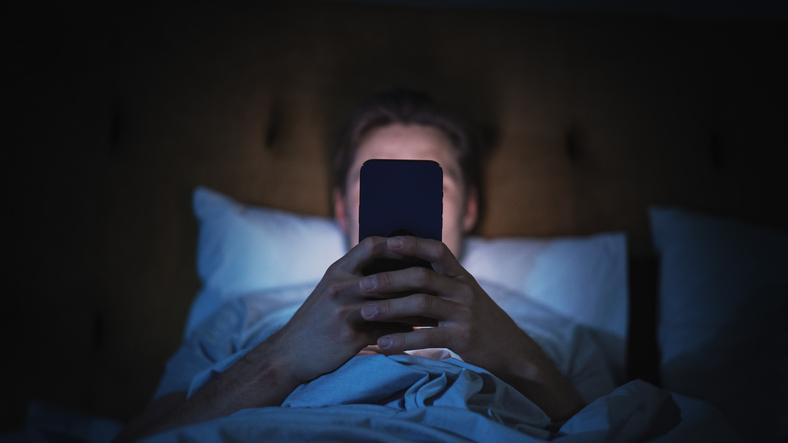 A man sits in darkness, illuminated by a smartphone that blocks his face from view
