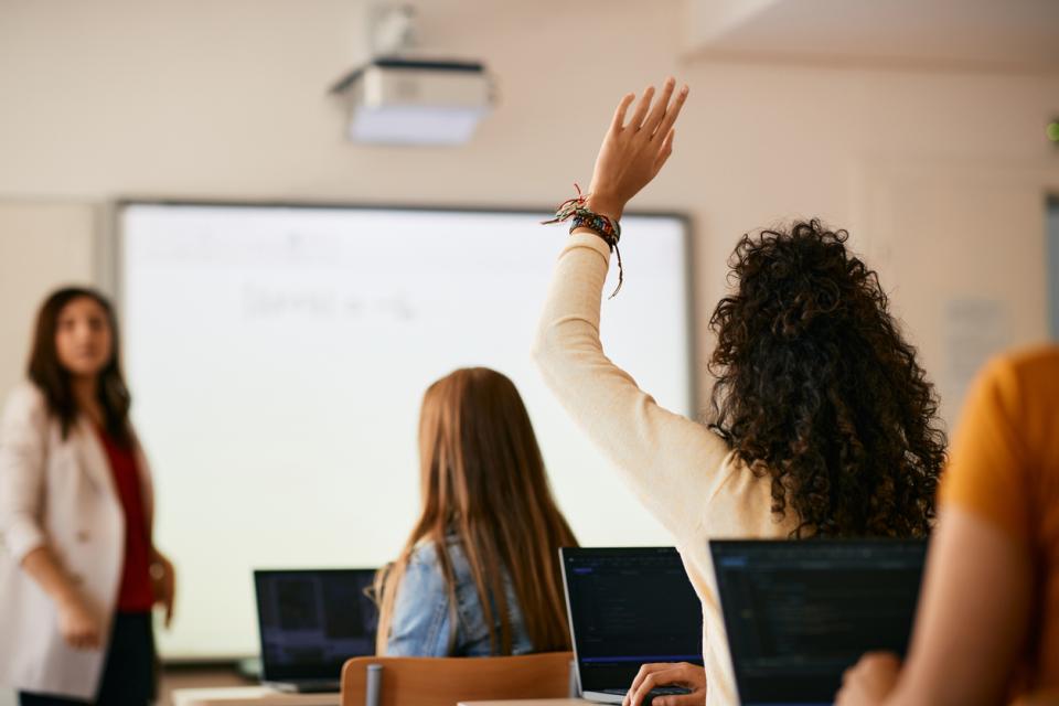 A class full of students from behind, with one raising their hand to answer a question