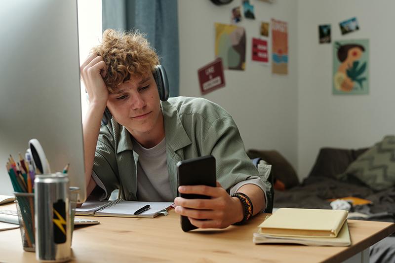 Young male student looking at phone while putting off study
