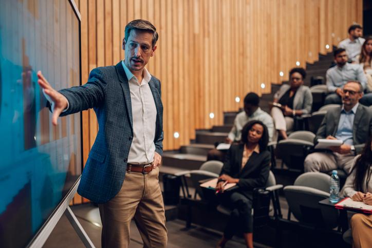A teacher gestures to a screen in a lecture hall