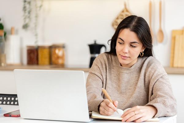 A student taking notes on a notepad while looking at her laptop