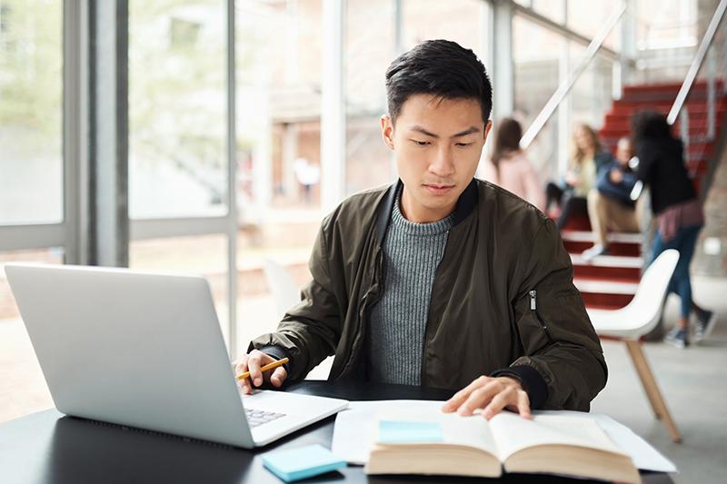 Male Asian student writing on laptop in common area