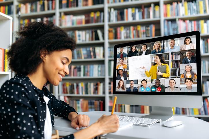 A female student taking notes from her online class with other students and a teacher appearing in a gallery view on her screen