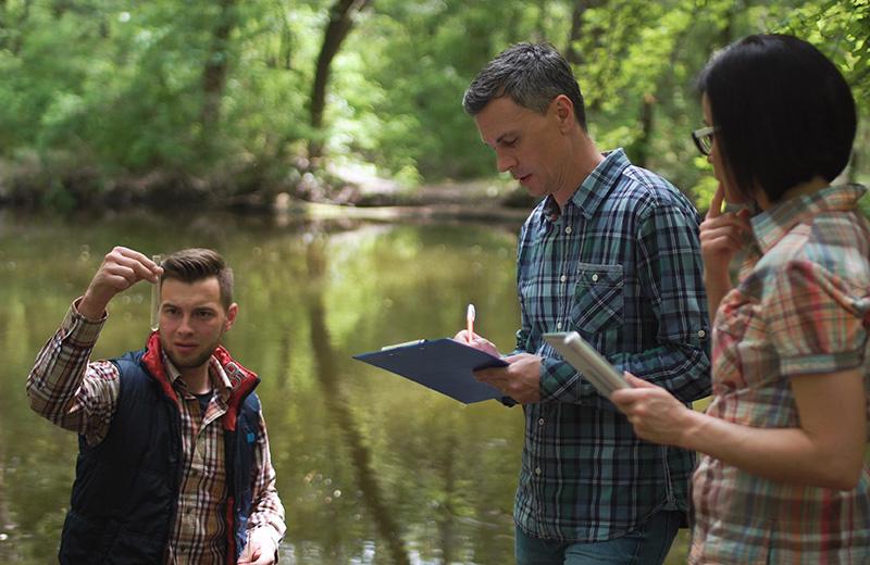 Three researchers in casual clothing at a lake take water sample