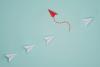 red paper airplane leaving line of white paper airplanes